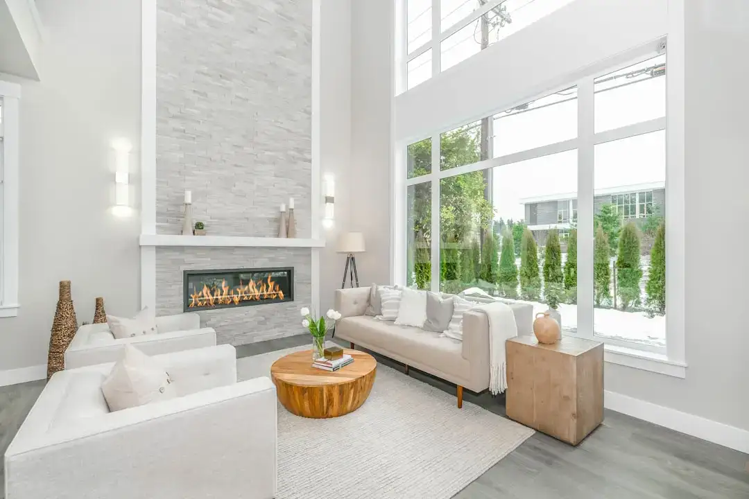 Spacious living room with large windows, white furniture, and a cozy fireplace, creating a bright an.